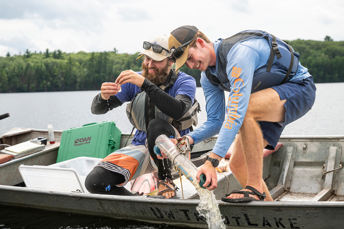 Two people kneel on an aluminum boat floating in a lake. One person empties a long cylinder of lake water back into the lake, while the other person behind him closes a small water sample.