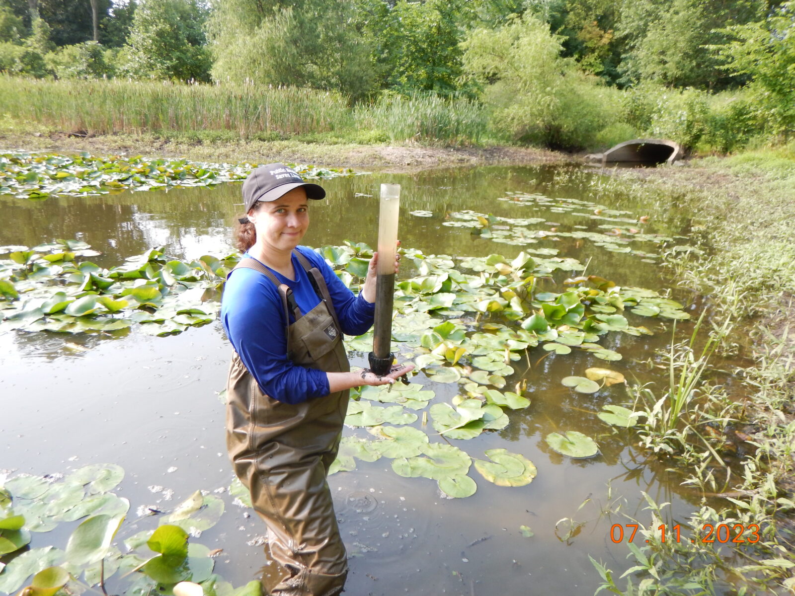 A person in waders stands in a pond holding a cylinder full of sediment.
